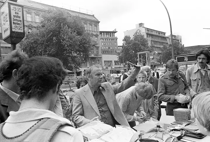 Straßenhändler preist seine Ware an, dahinter eine Litfaßsäule mit B.Z.-Reklame