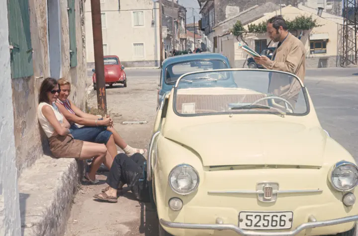 Panne mit einem cremefarbenen Fiat 600 Cabrio in einer Dorfstraße, zwei Frauen warten auf der Mauer, ein Mann liest die Karte, ein Monteur liegt unter dem Auto, Dia von 1971