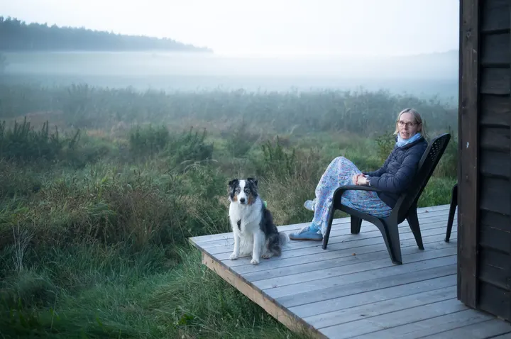 Frau im Gartenstuhl und Australian Shepherd auf einer Holzterrasse vor nebliger Wiese