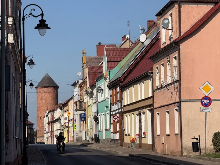 Straße mit bunten Stadthäusern und rundem Backsteinturm der Stadtmauer in Maszewo