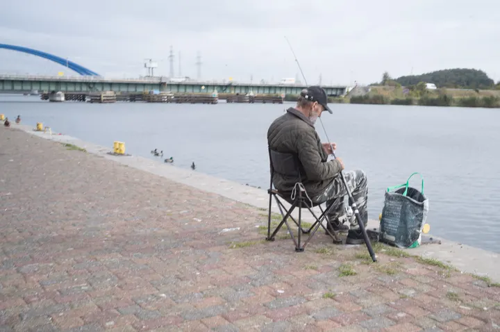 Angler auf einem Klappstuhl an der Dziwna, dahinter eine Brücke