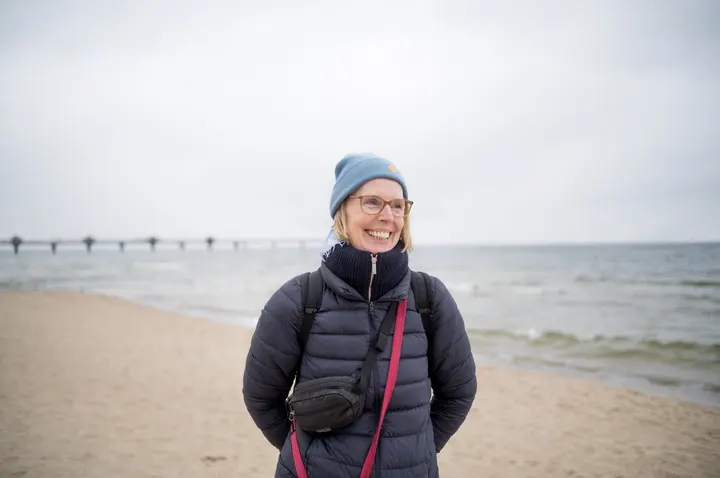Lachende Frau mit Mütze am Strand, im Hintergrund die Seebrücke