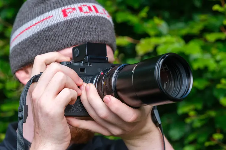 Fotograf mit Mütze blickt durch den Visoflex 2 der Leica M11, davor das lange APO-Telyt-R 180 mm