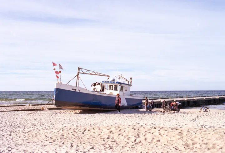 Blau-weißes Fischerboot am Ostseestrand, daneben Fahrräder und Spaziergänger